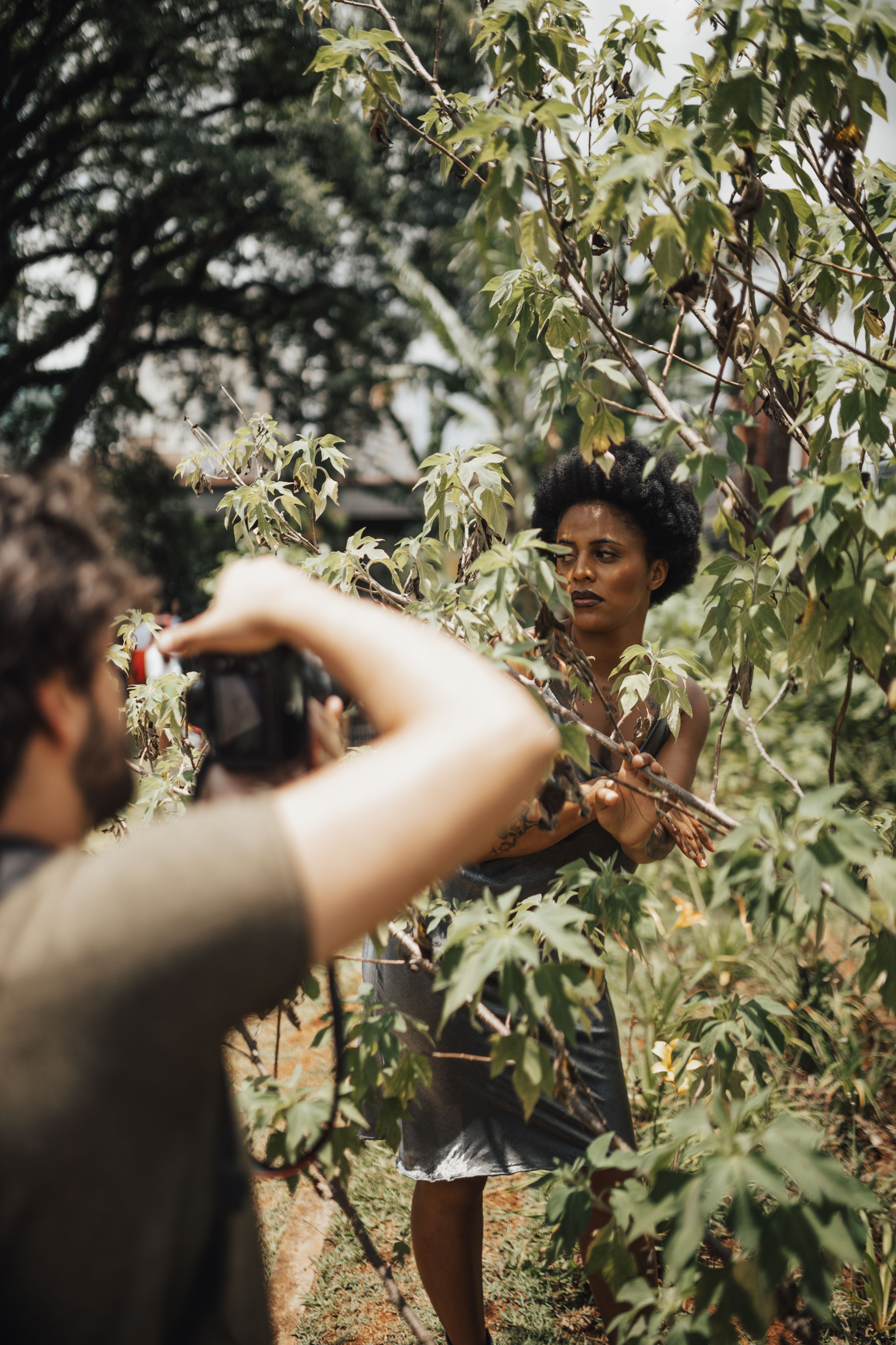 A man captures a woman posing among greenery during a sunny photoshoot.
