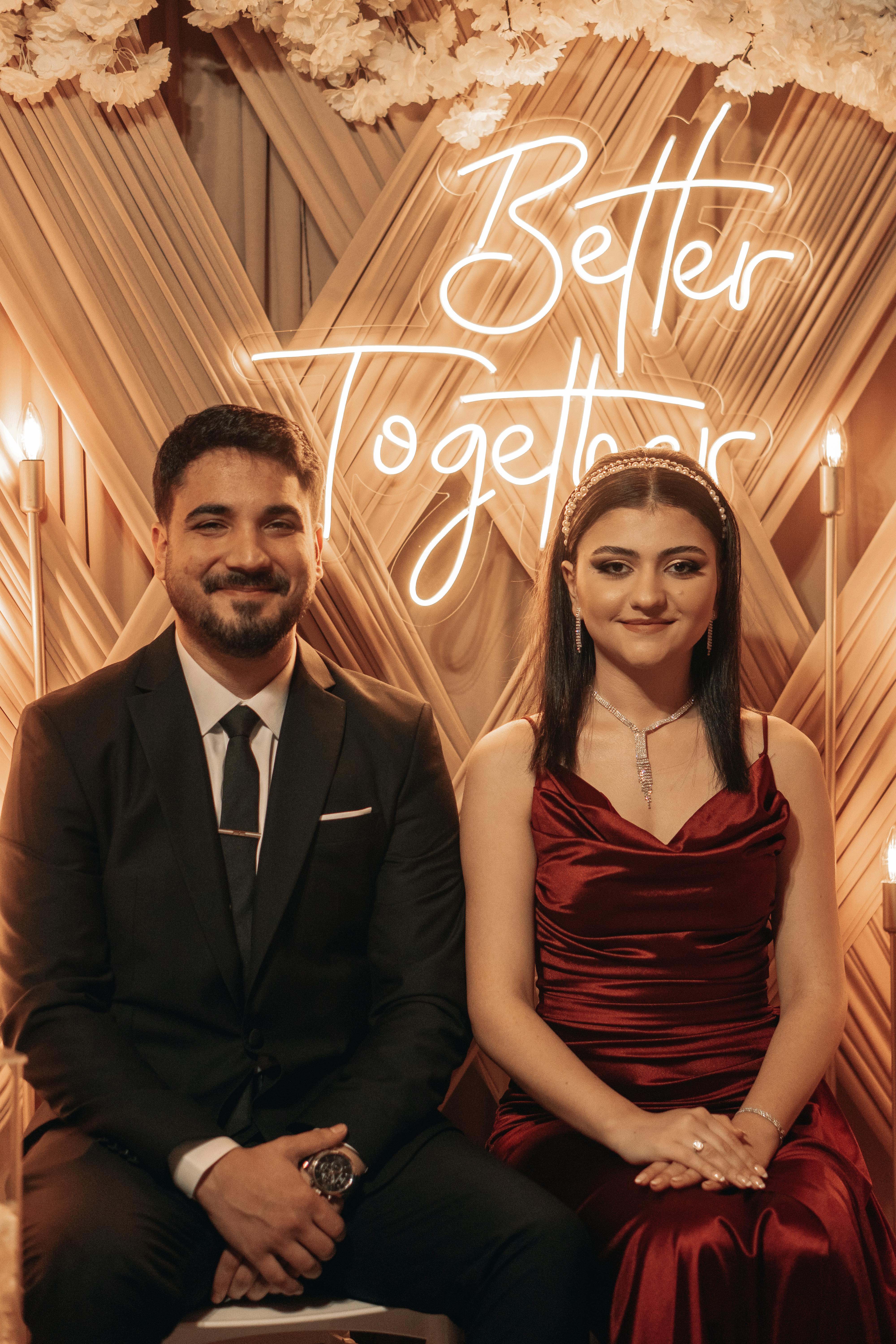 A smiling couple elegantly dressed, posing together at a celebration with a neon 'Better Together' sign.