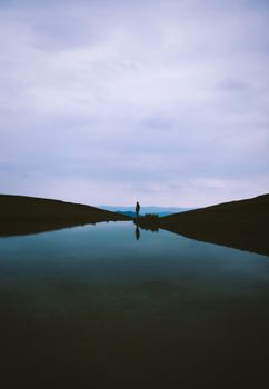 A calm and symmetrical landscape with a silhouetted figure reflecting in the water under an overcast sky.