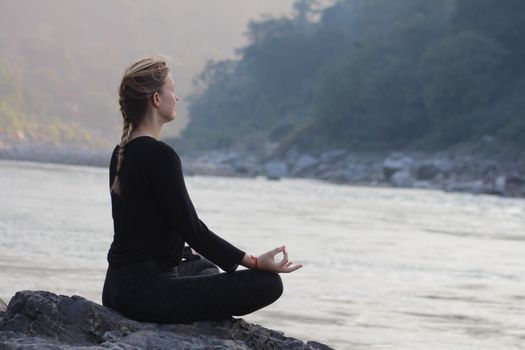 Peaceful scene of a woman meditating by the Ganges River in Rishikesh, India at sunrise.