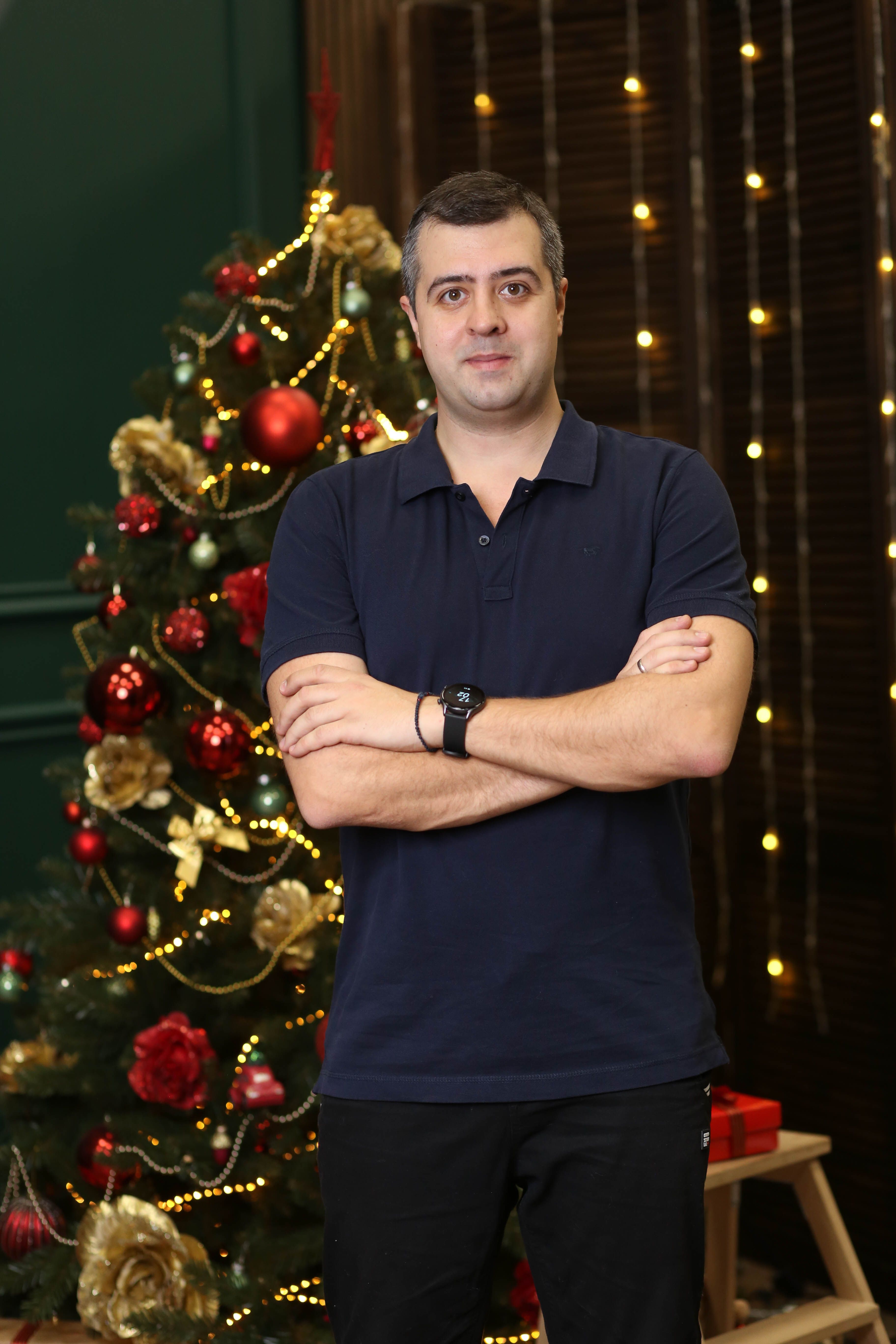 Adult man standing by illuminated Christmas tree, arms crossed, festive mood.
