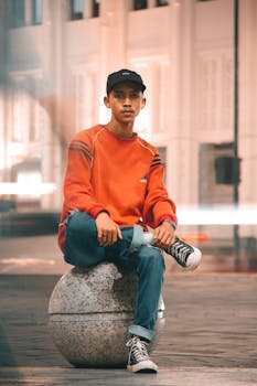 Stylish young man sitting on a stone ball in urban night setting wearing streetwear.