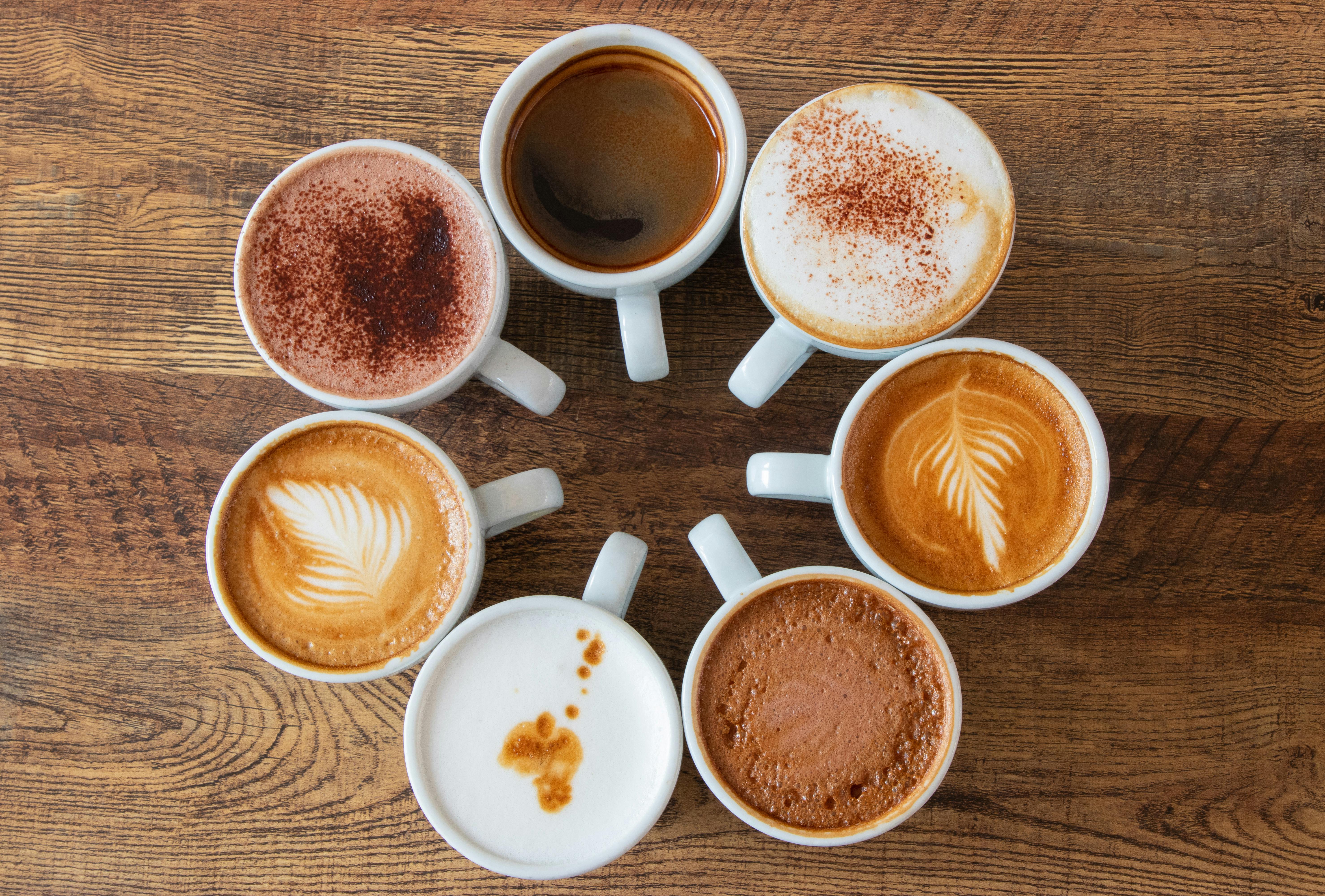 Top view of various coffee cups with latte art and foam arranged in a circle on a wooden table. What Is Cappuccino?