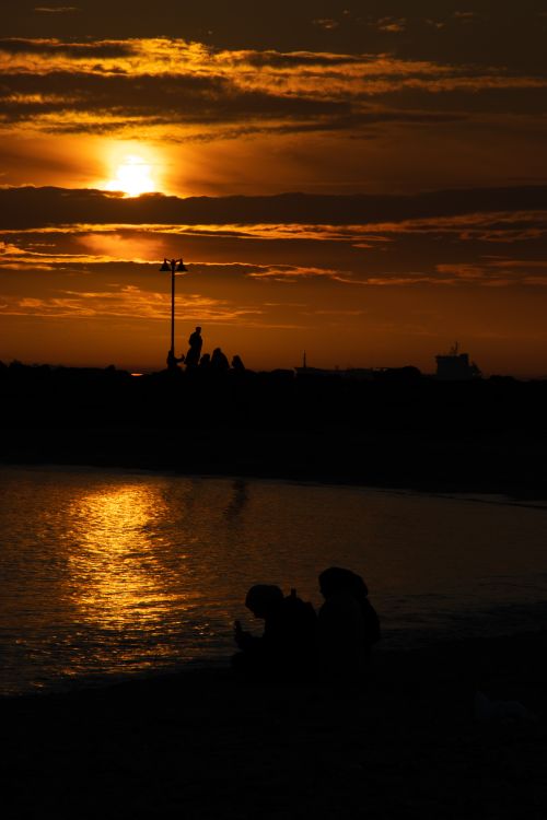 Free Silhouettes by the water during a dramatic sunset, reflecting warm hues. Stock Photo
