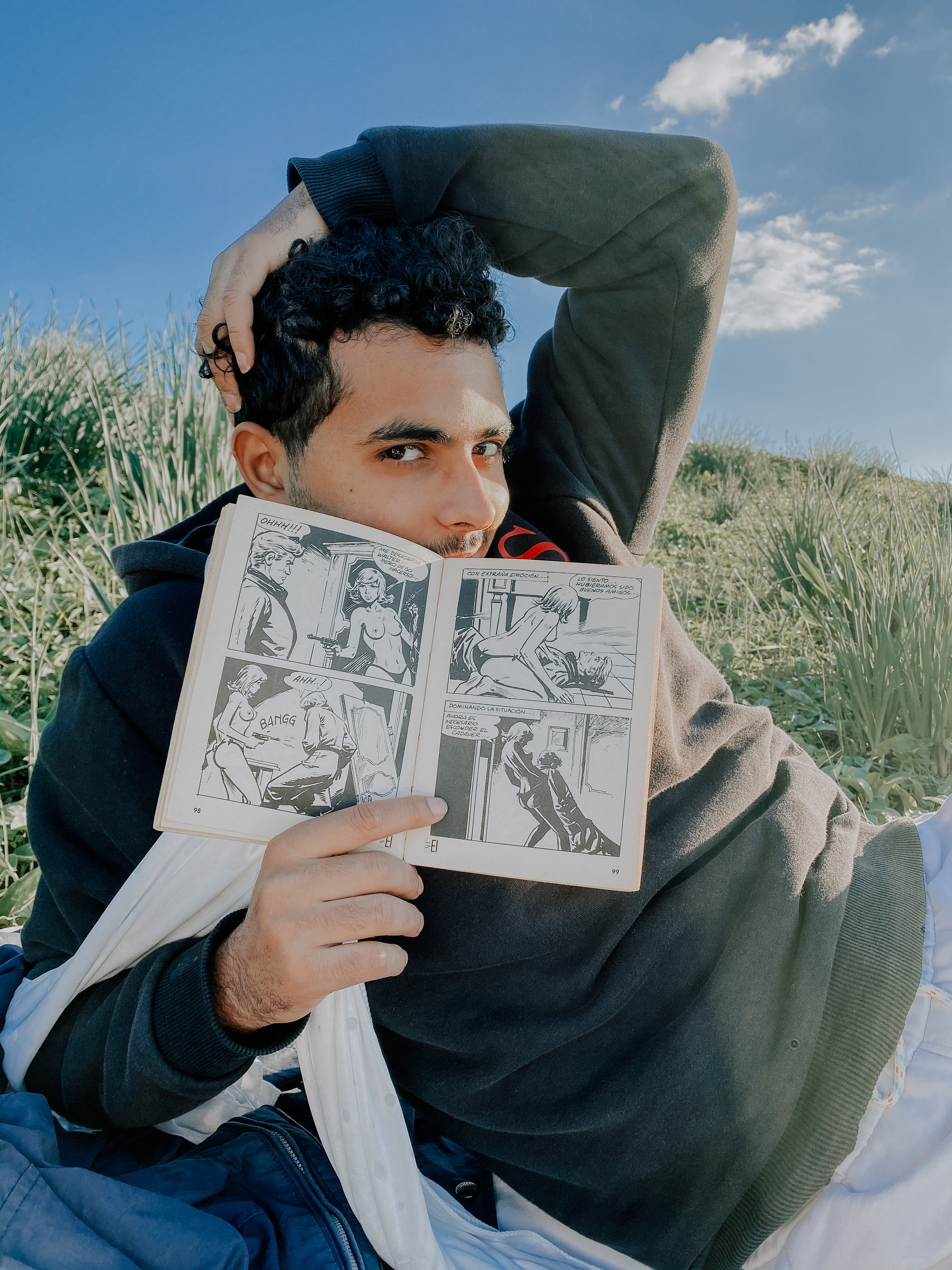 A young man with curly hair reads a comic book while relaxing in a grassy field under a clear blue sky.