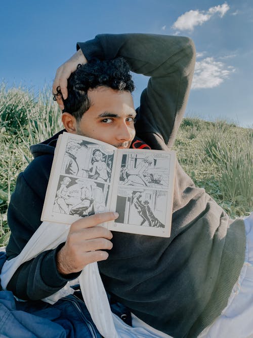 A Man in Hoodie Holding a Comic Book while Sitting on a Grass Field