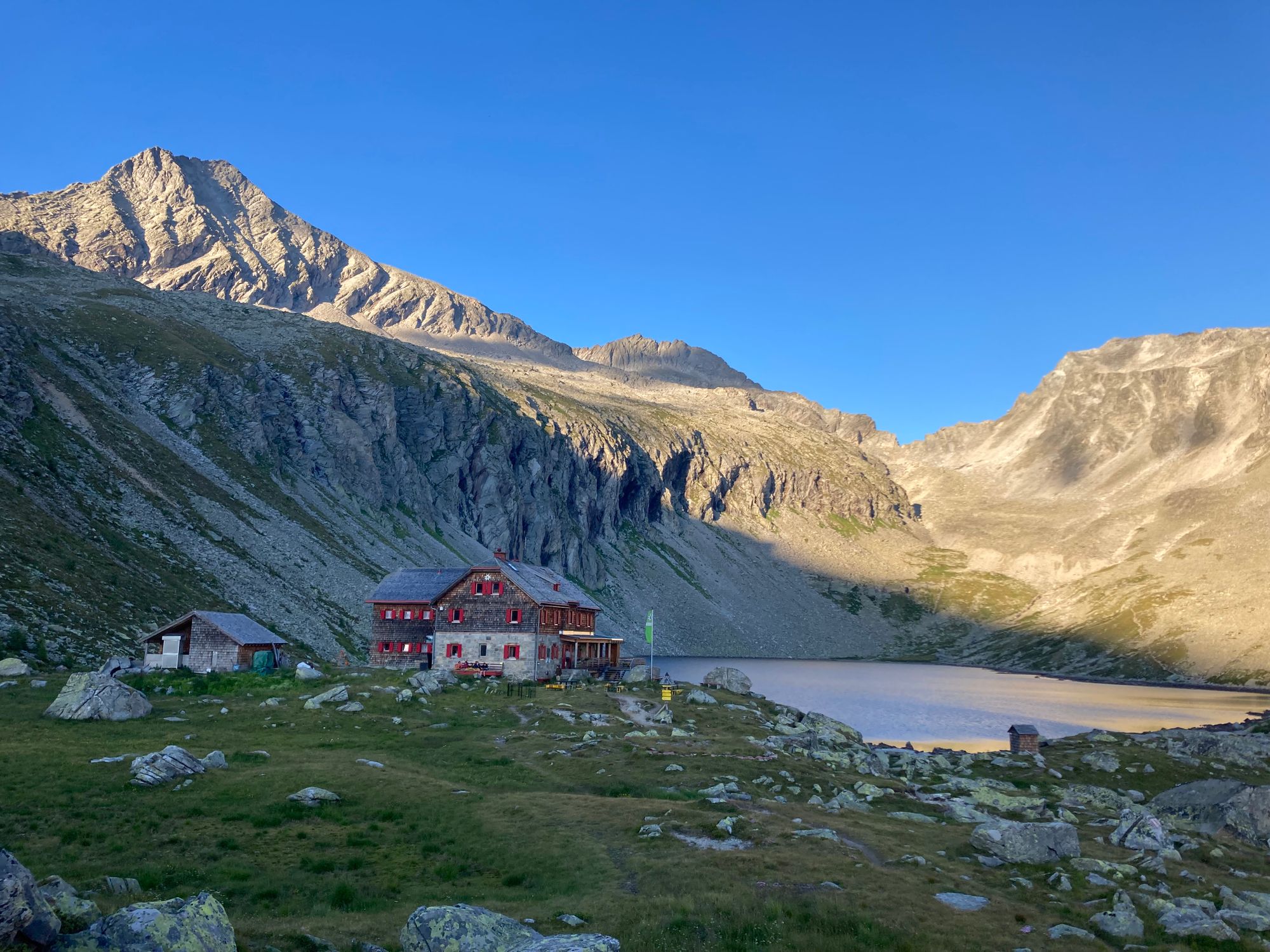 A mountain lodge nestled by a tranquil lake in a rugged alpine landscape under a clear blue sky.