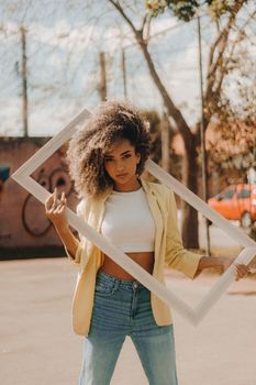 Confident woman with curly hair poses outside, holding a frame in an urban setting.