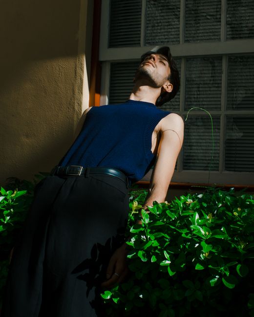 A stylish man in a sleeveless top poses leaning against a window, surrounded by fresh greenery.