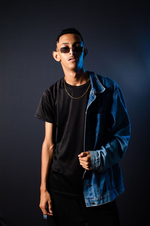 Free Young adult in dark studio wearing sunglasses and denim jacket, showcasing modern street style. Stock Photo