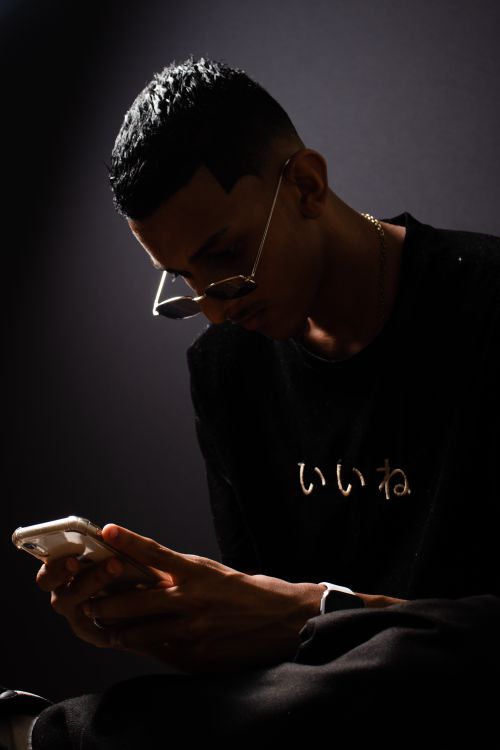 Free A young man wearing sunglasses and a black shirt texts on his smartphone in a dimly lit setting. Stock Photo