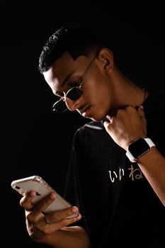 Portrait of a young man with sunglasses using a smartphone, wearing a smartwatch against a dark background.