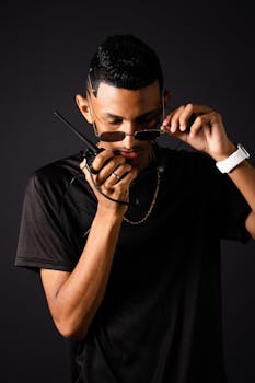 Man in sunglasses and black t-shirt holding walkie talkie against black background.