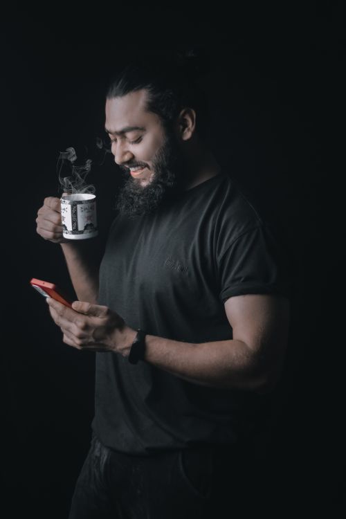 Free Smiling bearded man with a bun, holding coffee, using smartphone in studio setting. Stock Photo