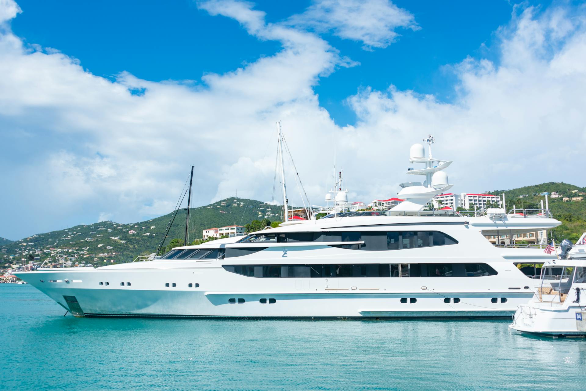 A sleek luxury yacht docked in a tropical harbor with clear blue skies and picturesque hills.