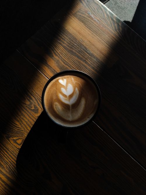Free A beautifully crafted latte with heart foam art sits on a dark wooden table with natural light and shadows. Stock Photo