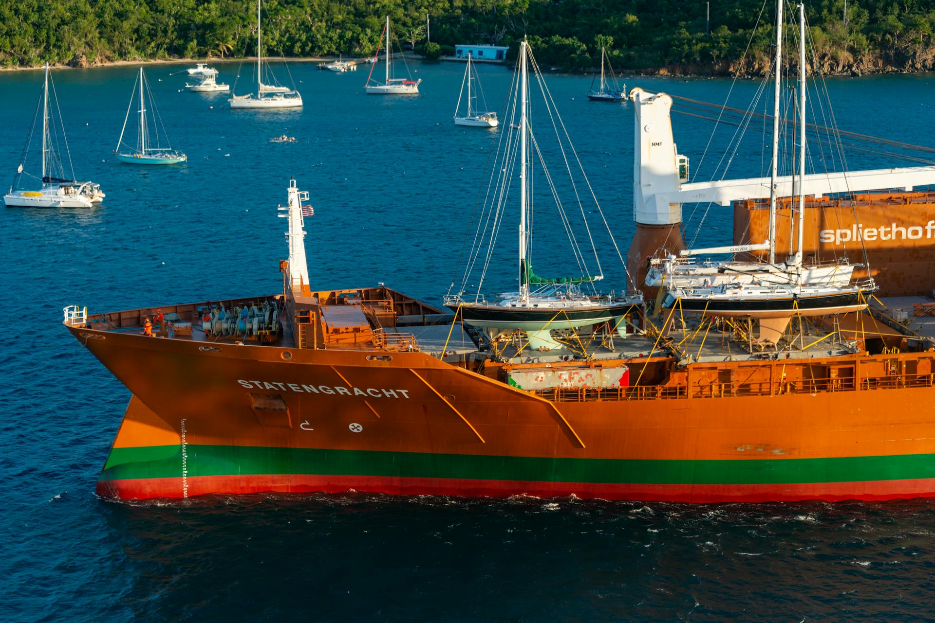 A vibrant cargo ship named Statengracht loaded with sailboats sailing in clear blue coastal waters.