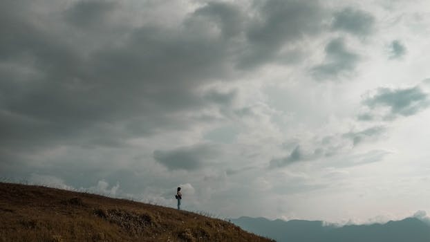A solitary traveler stands under a vast cloudy sky on a hillside in Matur, Indonesia.