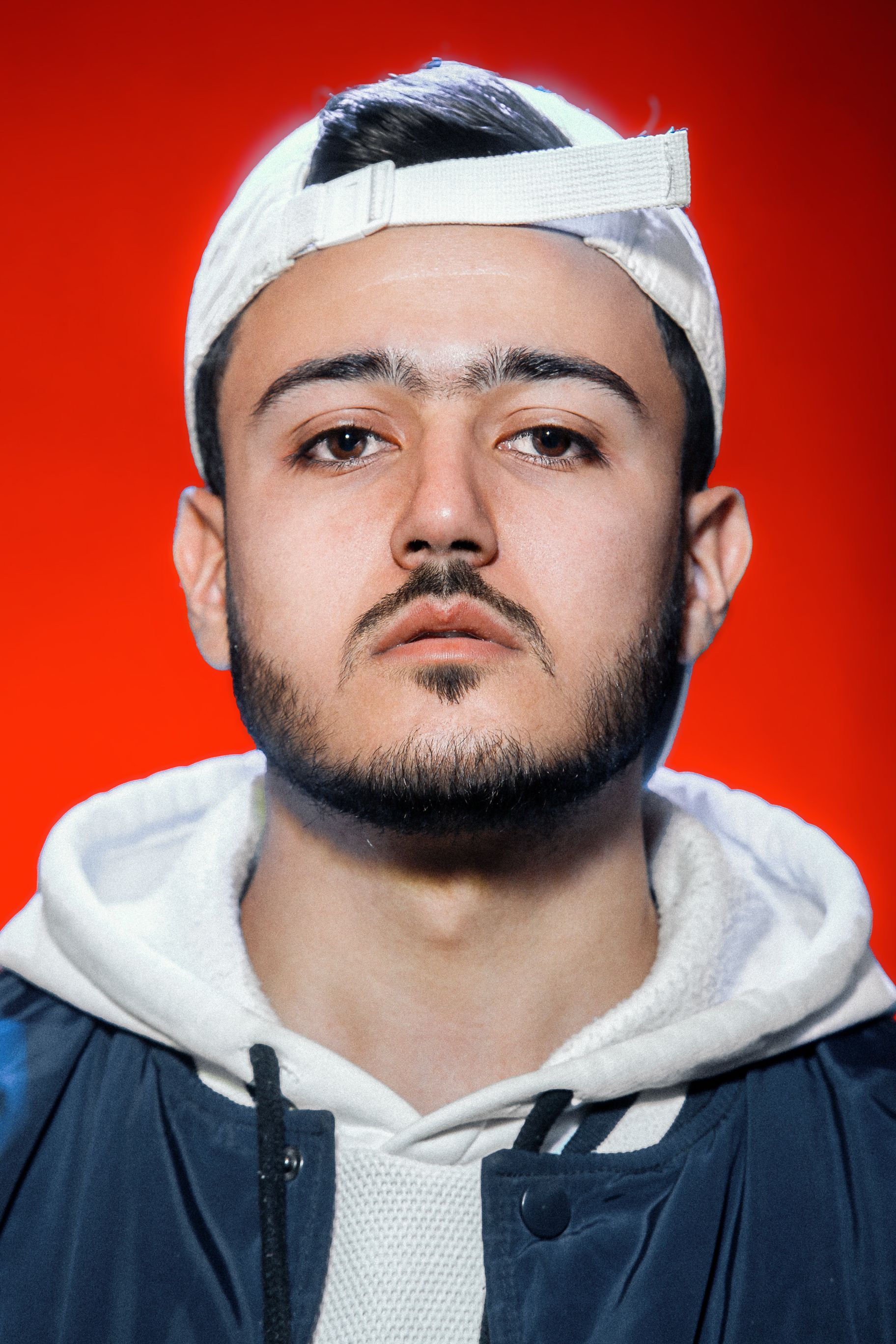 Fashion portrait of a young man wearing a white hoodie and cap against a red studio background.