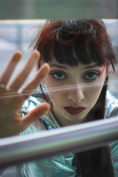 Free A young woman with dark makeup and bangs gazes seriously through a transparent glass surface. Stock Photo