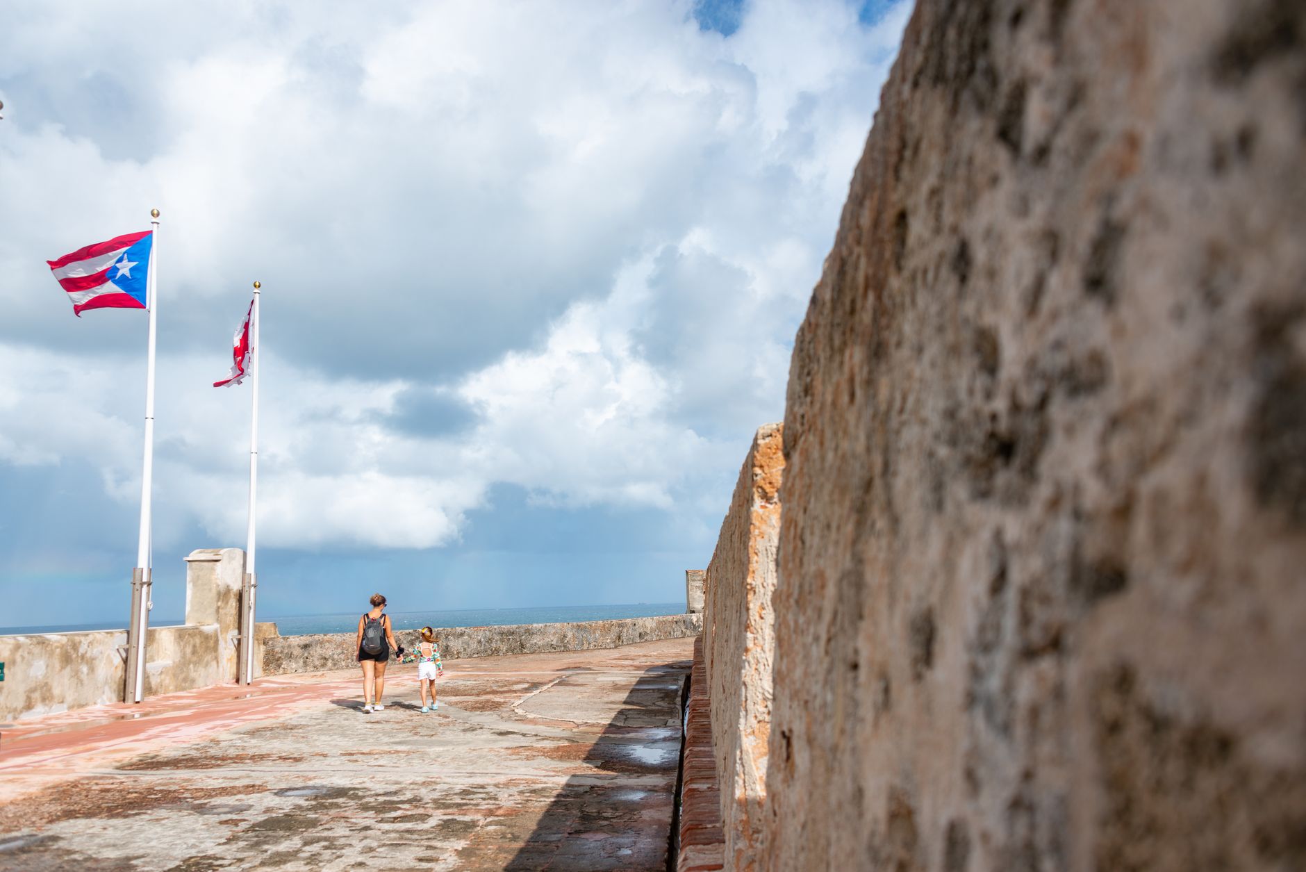 A mother and daughter walk along El Morro fortress with flags in the breeze, under a cloudy sky.