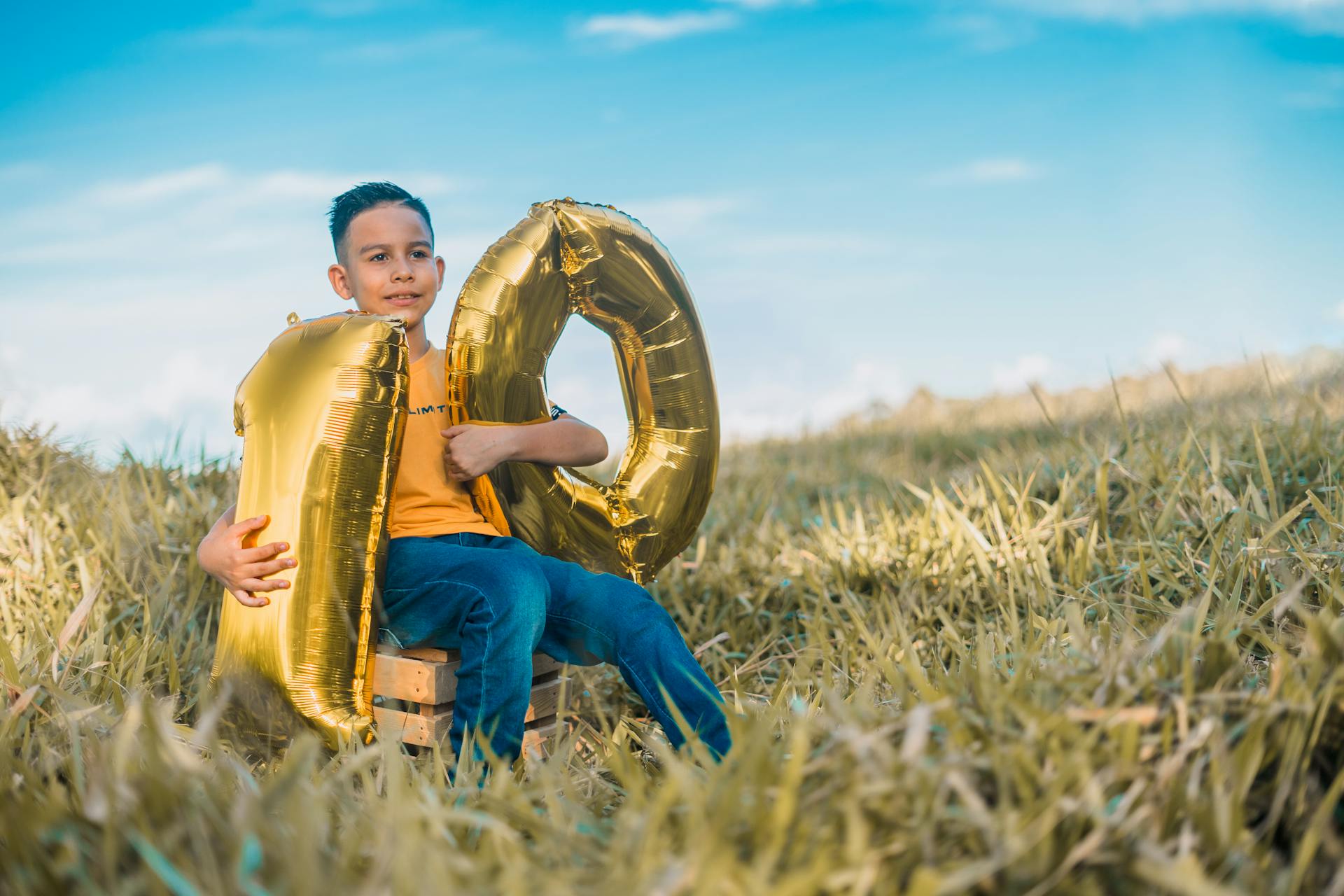 Un jeune garçon assis avec des ballons dorés formant un " 10 " dans une prairie ensoleillée, célébrant son anniversaire.