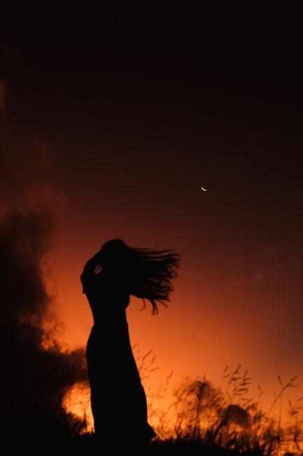 Silhouette of a woman with flowing hair at twilight against a crescent moon and orange sky.
