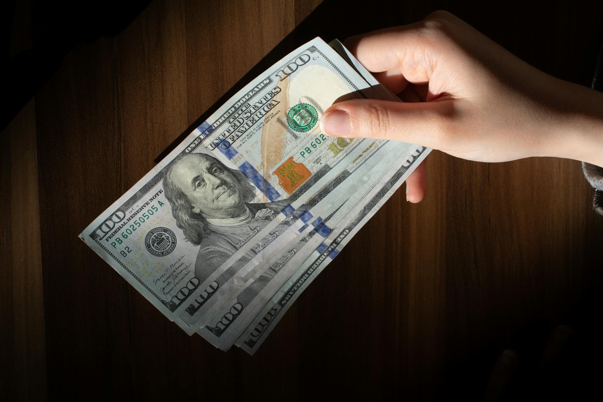 A hand holding a stack of 100 dollar bills on a wooden table, highlighting currency wealth.