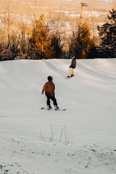 Two people snowboarding on a snowy slope surrounded by trees during wintertime, creating a leisure activity scene.