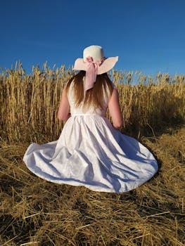A woman in a stylish white dress and hat sits in a golden wheat field under a clear blue sky.