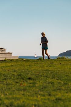 Woman jogging on a coastal path with a sailboat in the background, capturing a serene outdoor activity moment.