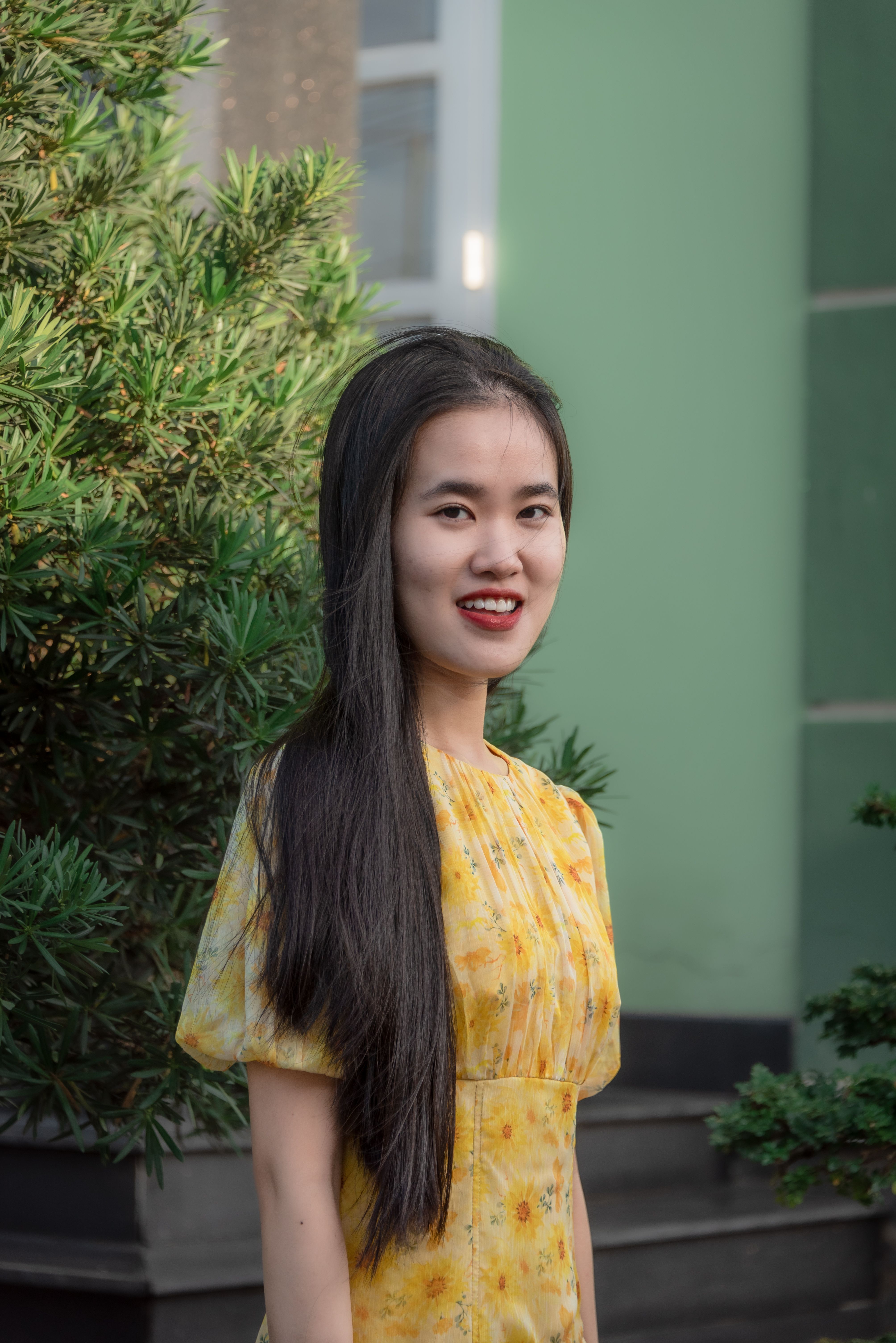 Young Asian woman with long hair in a yellow floral dress smiling outdoors on a sunny day.