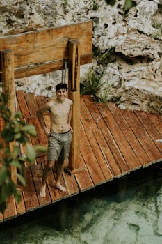 A smiling young man stands shirtless on a wooden boardwalk by a natural water pool, showcasing a relaxed summer vibe.