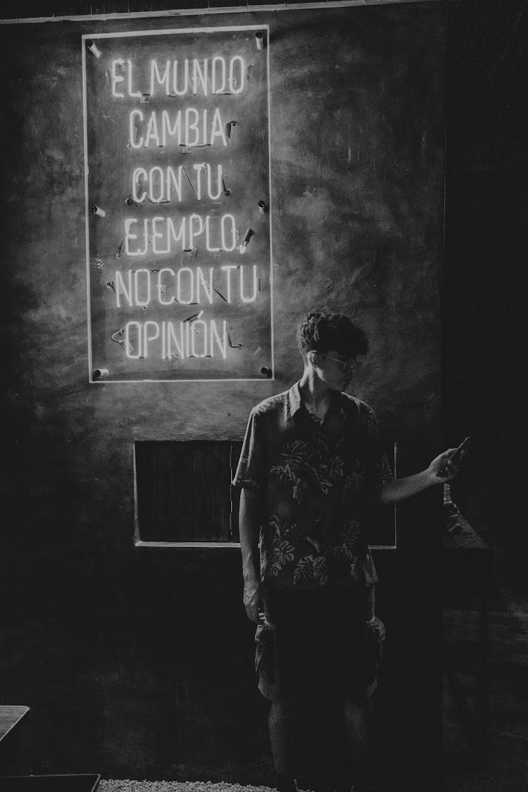 Black And White Shot Of A Man Standing In Front Of A Building With A Sign Behind 