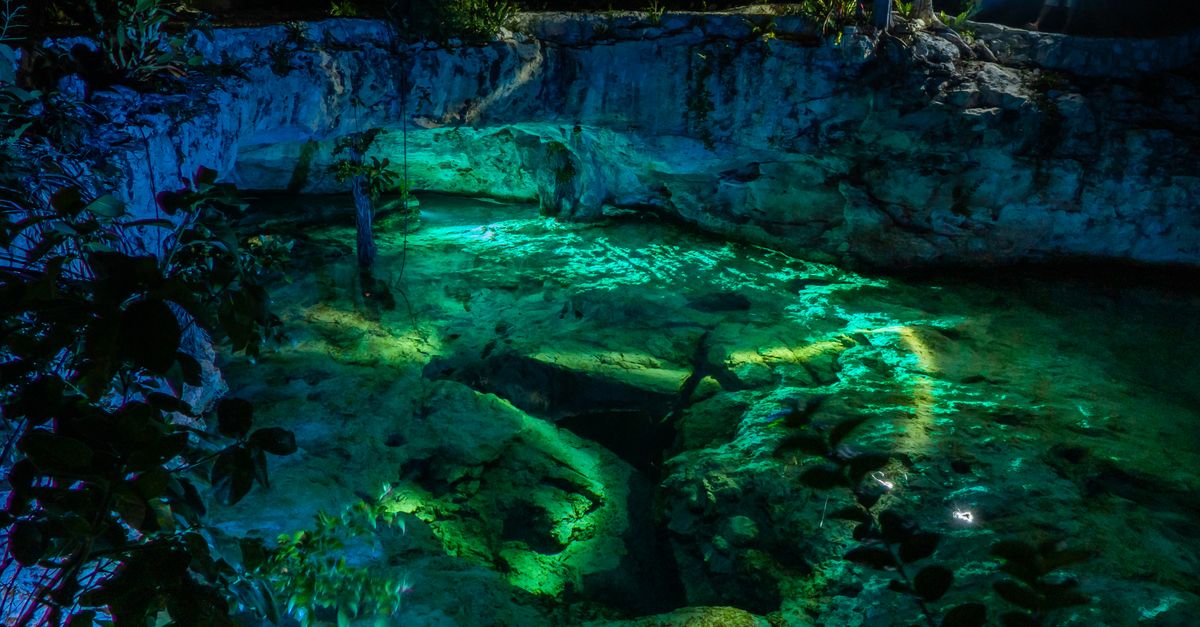 Vibrant night view of Grand Cenote in Tulum, Mexico, highlighting illuminated turquoise waters and rocky surroundings.
