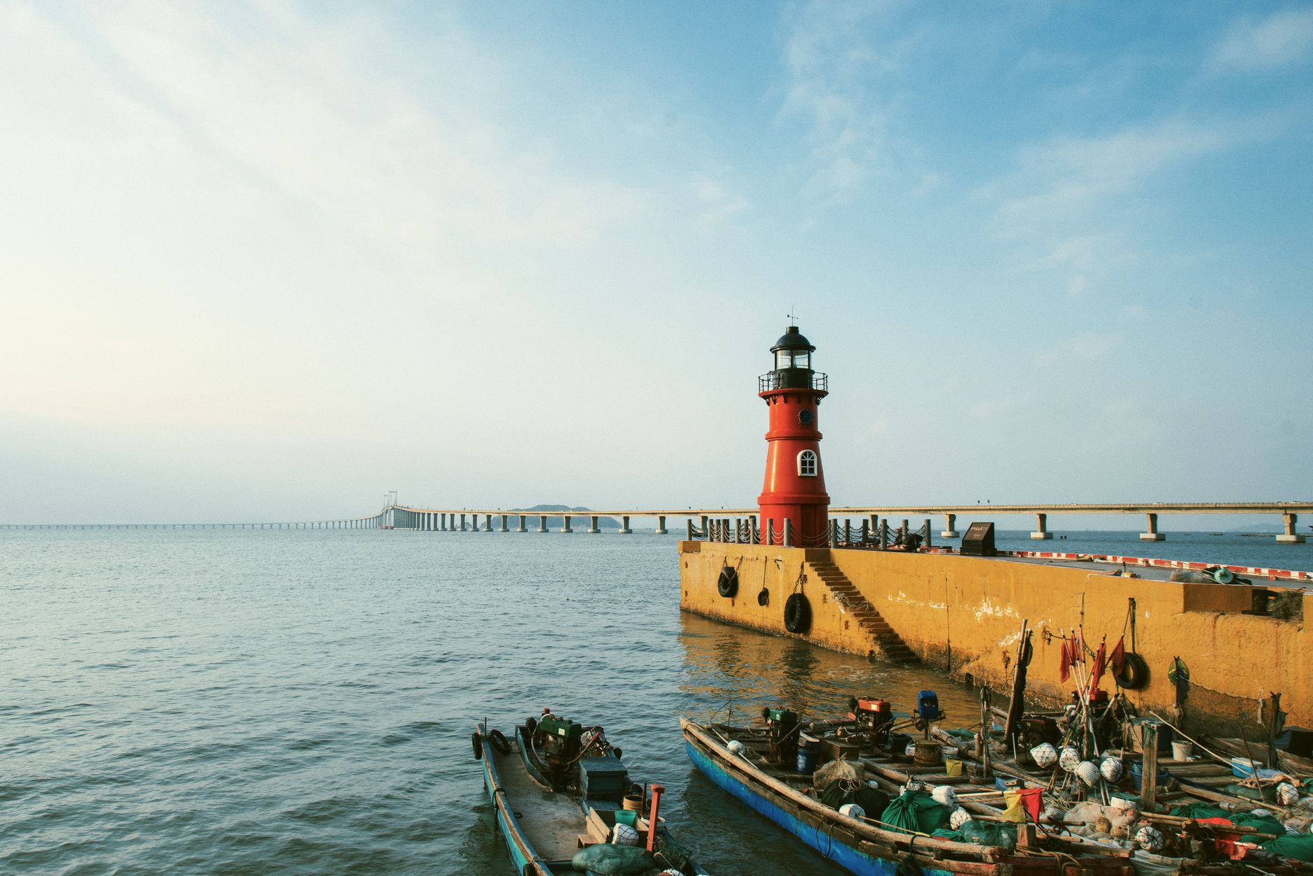 A scenic view of a red lighthouse on the seashore with boats and a pier stretching into the sea.