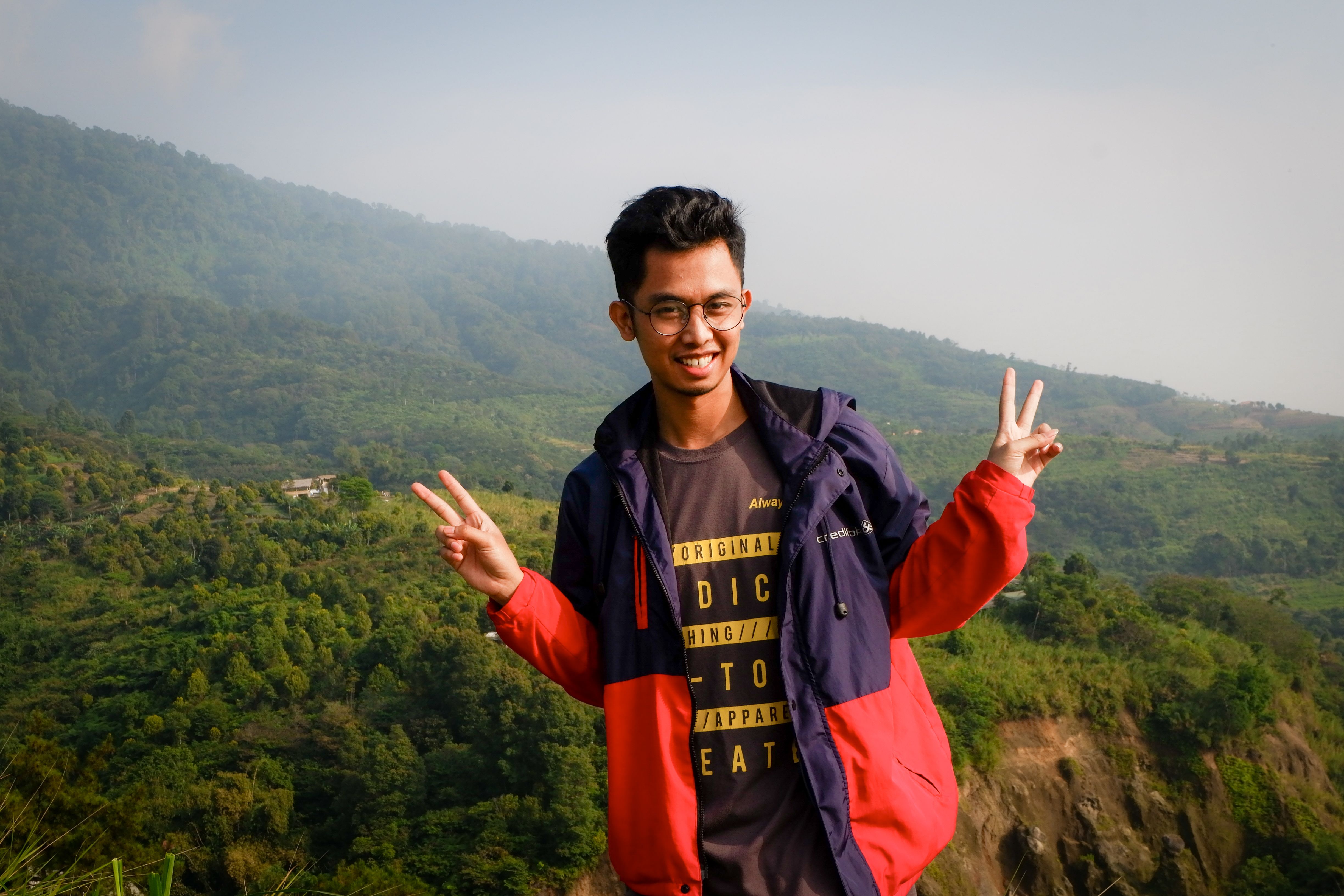 Asian man smiling with peace sign in lush green outdoor setting.