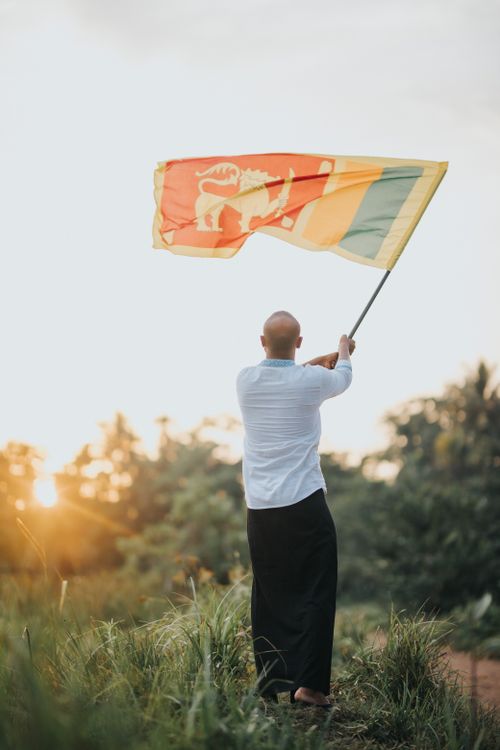 Free A man in traditional attire waves the Sri Lankan flag in a serene outdoor setting at sunset. Stock Photo