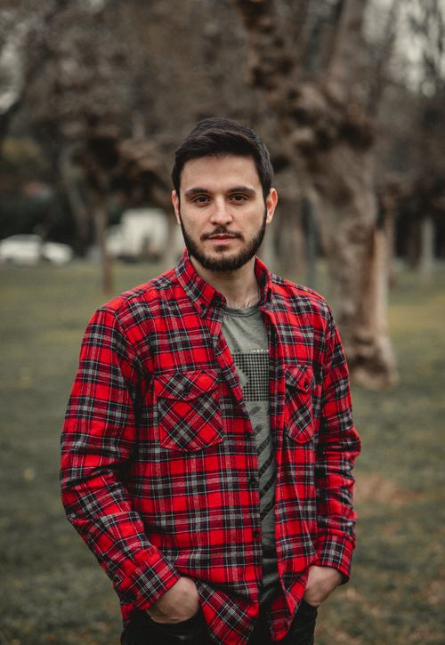 Free A bearded man in a red plaid shirt standing outdoors with hands in pockets. Stock Photo
