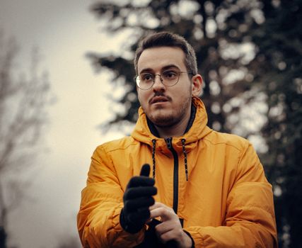 Portrait of a young man in a yellow jacket and glasses, standing outdoors with trees in the background.