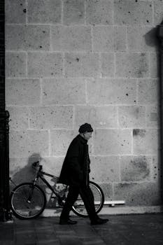 Monochrome image of a man walking past a bicycle against a city wall.