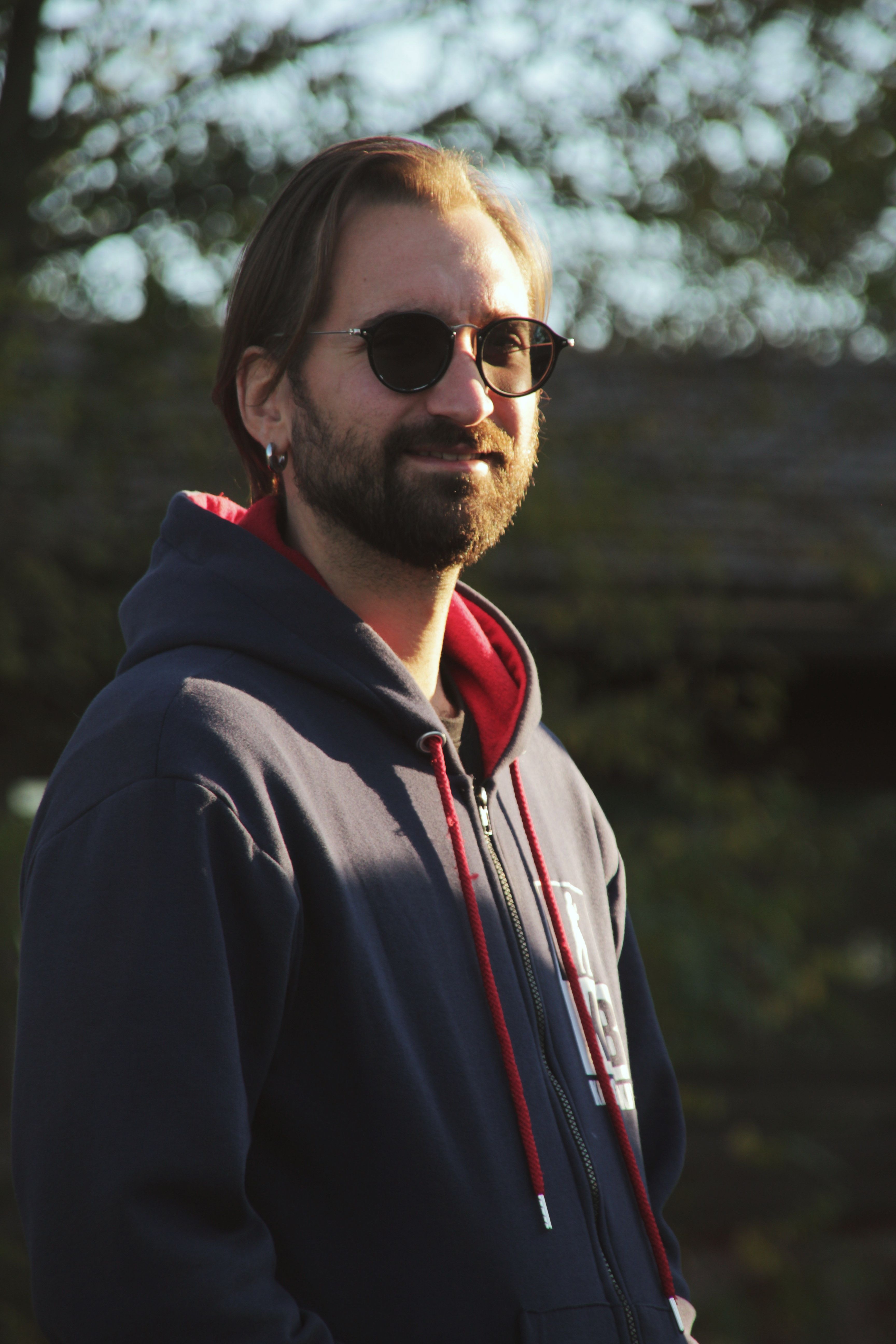 Portrait of a bearded man with sunglasses wearing a hoodie outdoors in a natural setting.