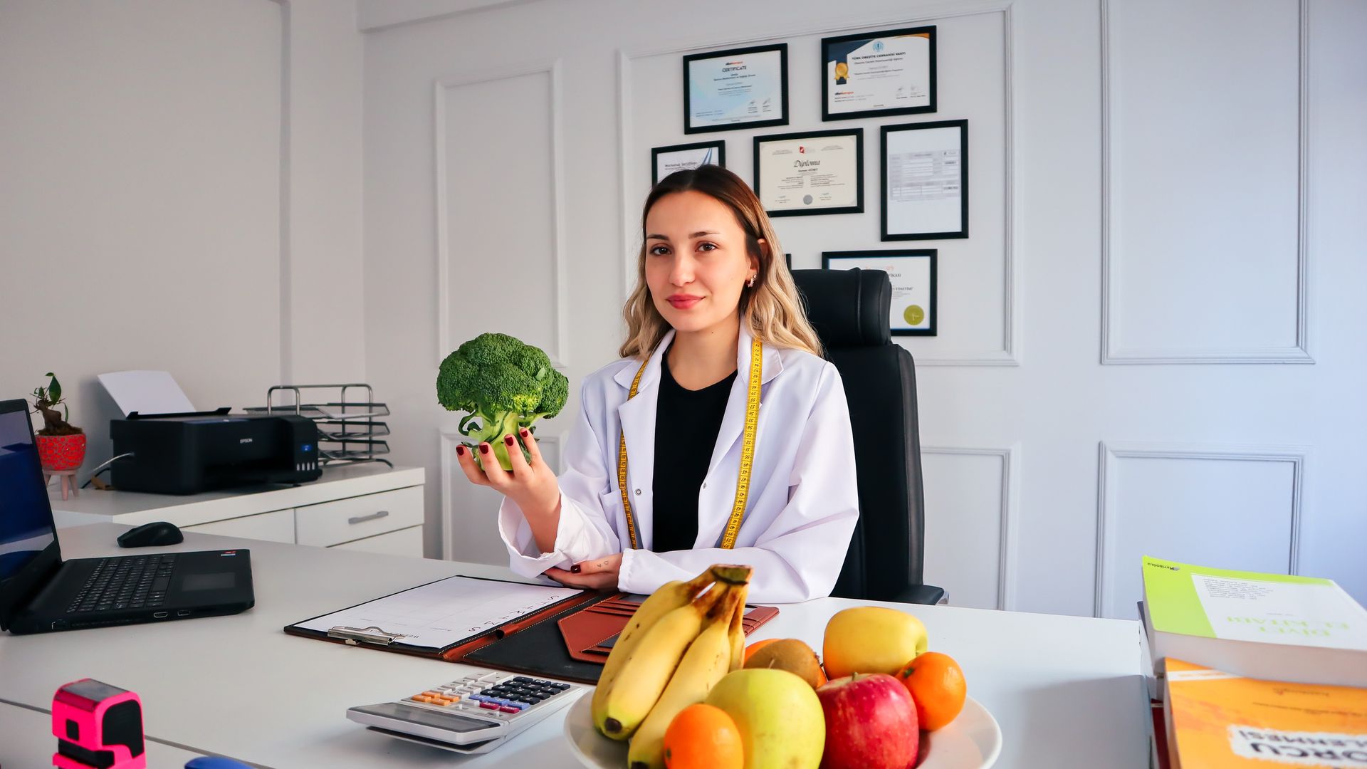 Female nutritionist in office holding broccoli, surrounded by fruits, promoting healthy lifestyle — how to health