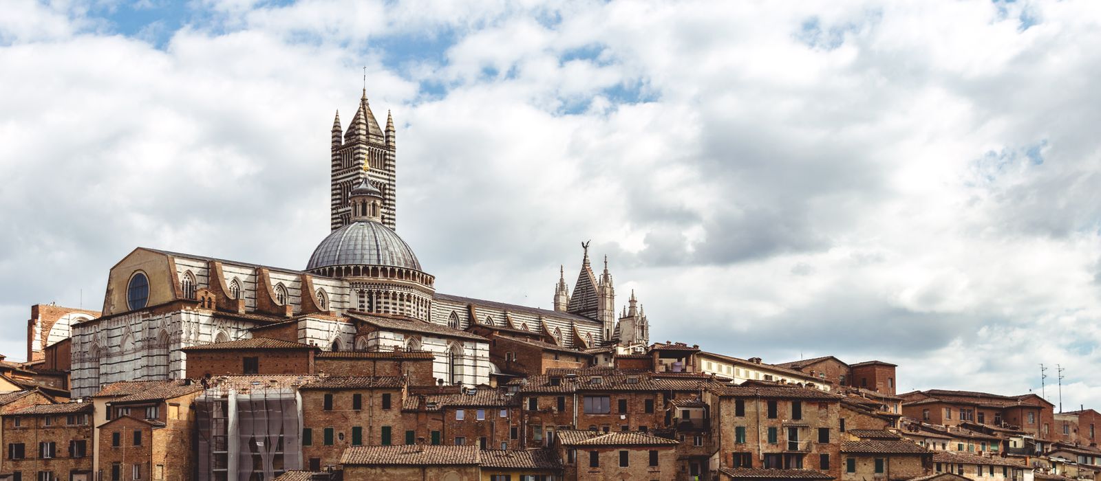 A scenic view of Siena's historic cityscape featuring the iconic Siena Cathedral under a cloudy sky in Tuscany, Italy.