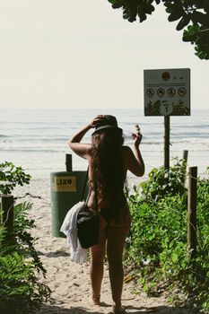 Woman in swimsuit walks towards Ubatuba beach, Brazil, through lush plants and sandy path.