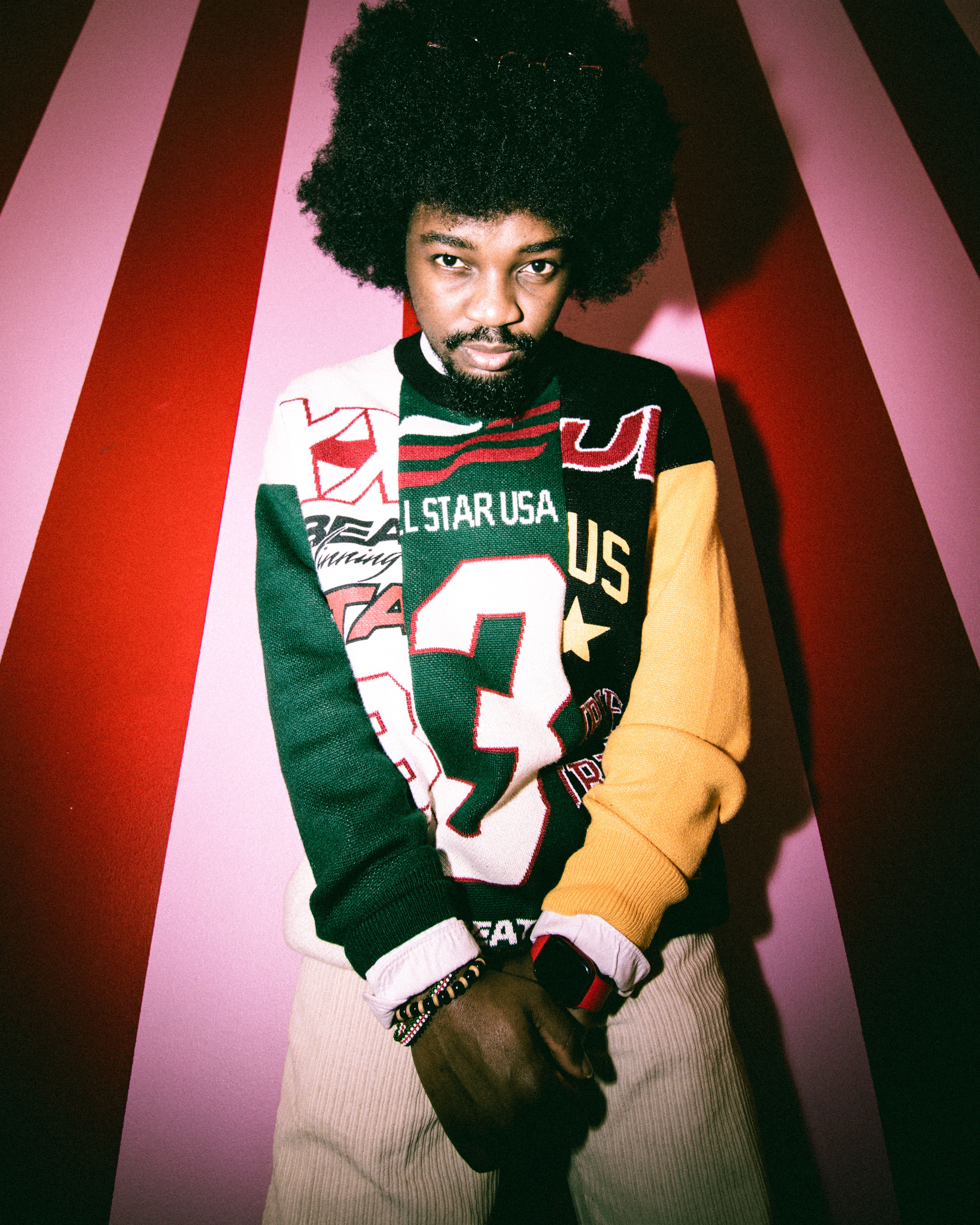 A man stands confidently in a colorful retro sweater against a vibrant striped background.