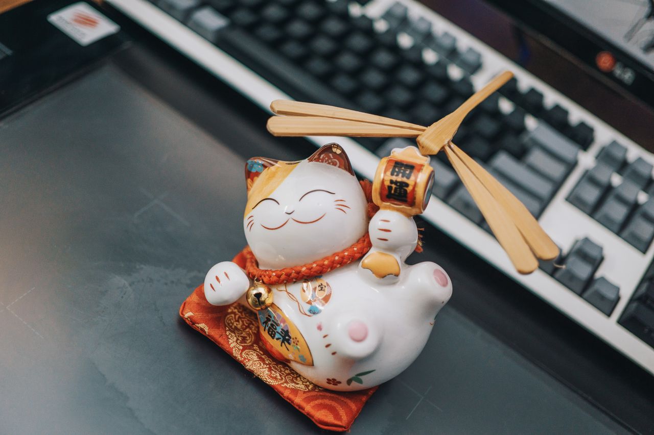 a ceramic maneki neko figurine sitting on a desk beside a keyboard sym