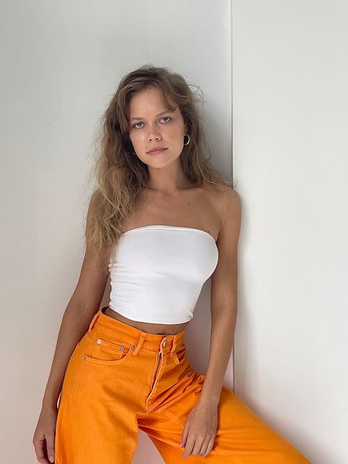 A Woman in White Tube Top and Orange Pants Leaning on a White Wall