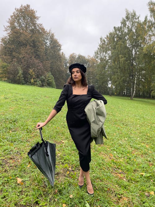 A Woman in Black Dress Holding an Umbrella while Standing on a Grass Field