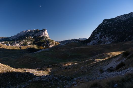 A stunning view of a rocky mountain landscape under a clear blue sky with a visible moon.